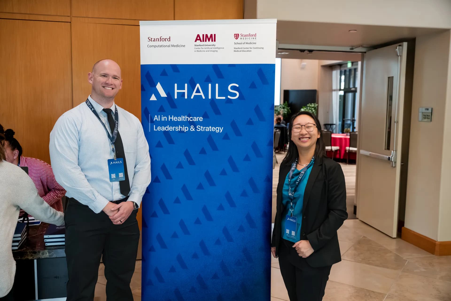 Two attendees standing either side of the HAILS banner, which carries the Stanford Computational Medicine, Stanford AIMI and Stanford Medicine marks